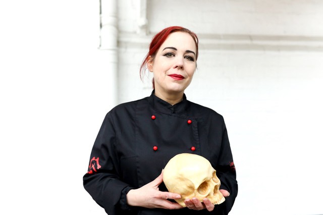 Annabel de Vetten with red hair, wearing a black chef&#039;s jacket with red buttons, and holding a skull