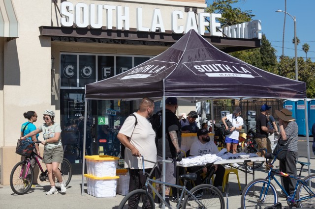 A crowd of bicyclists stopping by a pop-up tent for refreshments in front of a building with the sign &quot;SOUTH LA CAFE&quot;.