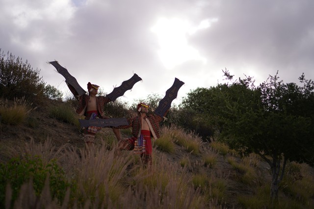 Two dancers on a grassy hill wearing red pants and decorative pieces tied to their arms