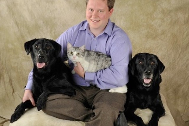 Dr. Christopher Schneider in a violet button-down shirt and holding a gray and white cat, sitting between two black dogs