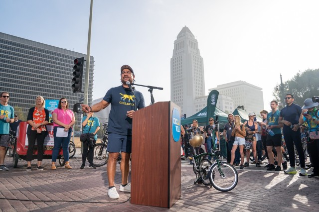 Man wearing a &quot;CicLAvia&quot; and P-22 t-shirt, speaking behind a podium, with a bicycle parked next to him, surrounded by a crowd of attendees, with L.A. City Hall in the distance.