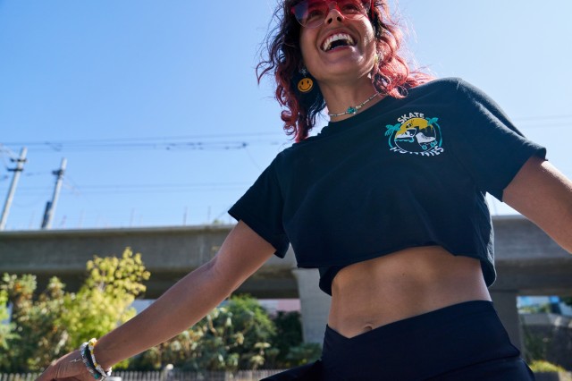 Woman with long pink hair laughing out loud, wearing a dark skirt and crop top with the logo &quot;Skate Hunnies&quot; on it.