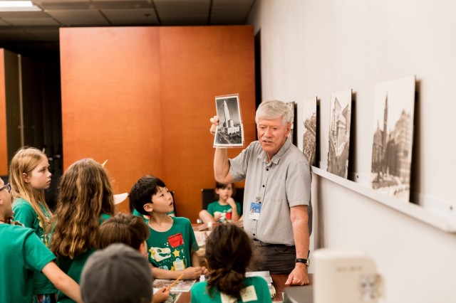 John Cahoon holding up a black and white photograph of a building towards a group of children in green t-shirts.