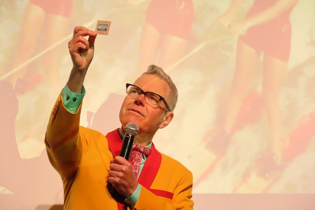 Charles Phoenix wearing an orange jacket with a red collar, holding up a photo slide, against a faded image of women&#039;s legs in red shorts on red skis