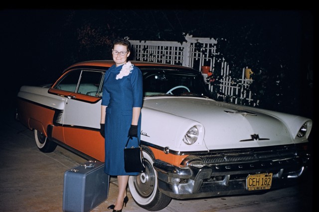Woman in a blue skirt next to a blue suitcase on the ground, leaning against an orange and white car