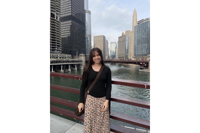 Christina Ela wearing a black sweater and flowered skirt with water and buildings in the background