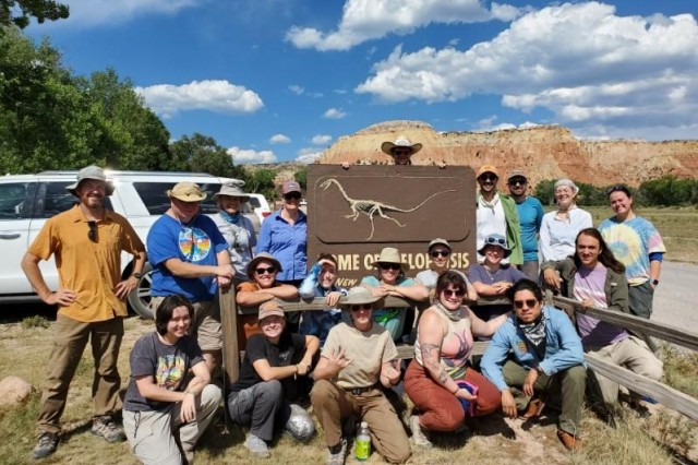 Group of excavators, some wearing tan hats, gathered around a sign with a dinosaur skeleton, with orange mountains and a blue cloudy sky in the background