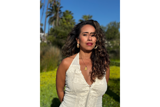 Emily Baeza with long, dark curly hair, wearing a white halter top, with her arms behind her back, and green grass, palm trees, and a blue sky in the background.