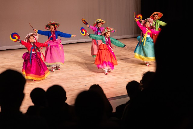 Silhouettes of audience members watching children in colorful outfits dancing on a stage while holding fans