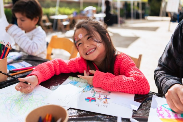 Young Asian-American girl smiling over her colorful drawing at an outdoor arts and crafts table.