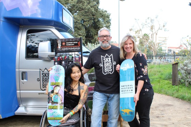 Two woman holding skateboards stand beside a man in front of a mobile skate shop.