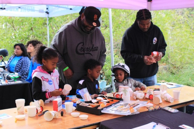 Two men stand behind three young children making crafts at a table.