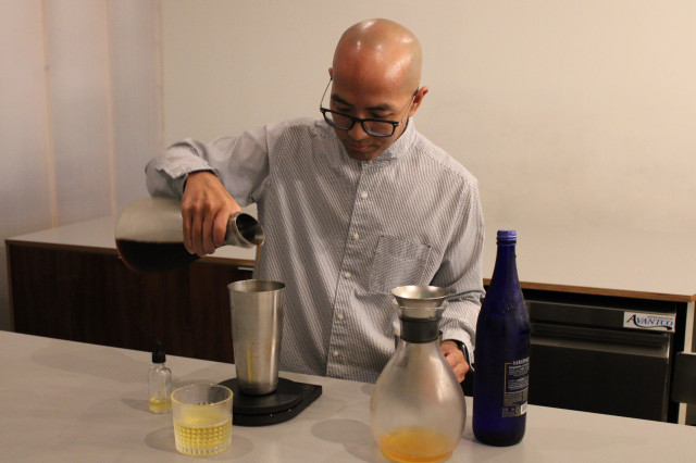 Bald man with black-rimmed glasses behind a minimalist bar, mixing a coffee cocktail.