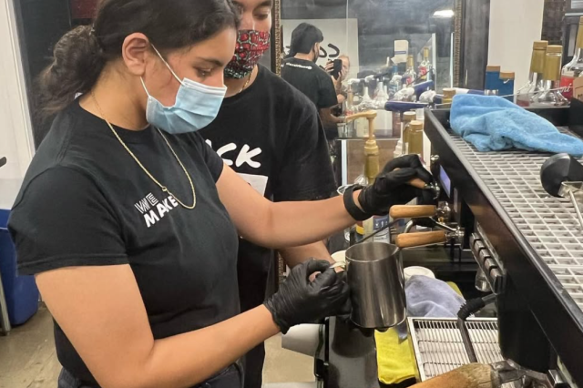 Two baristas dressed in black, wearing medical face masks, steaming milk at an espresso machine.