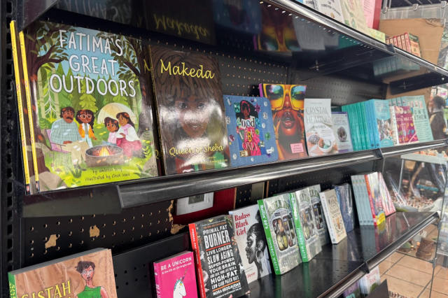 A close-up of a black bookshelf inside Simply Wholesome featuring books such as "Fatima’s Great Outdoors," "Makeda: Queen of Sheba," and "Primal Fat Burner."