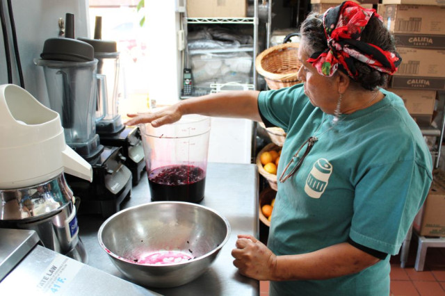 Woman in teal shirt with hand over large container with fruit inside, in kitchen