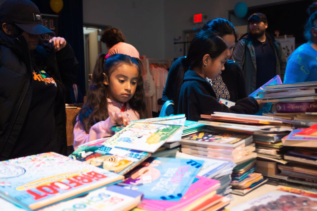  A young girl with brown hair and a pink sweater browses comics amidst the piles of books and comics at the CALE comic giveaway table