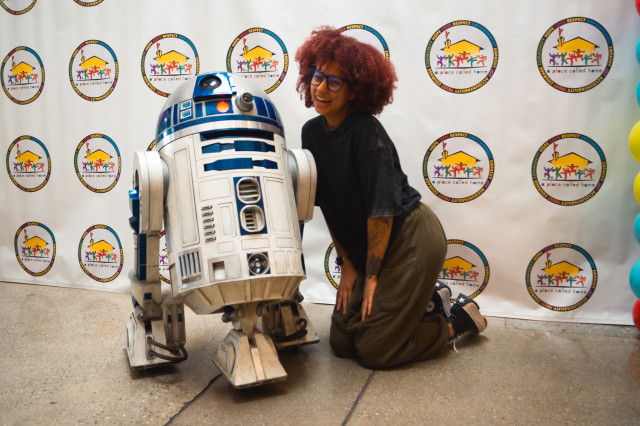 A red haired young woman poses, kneeling and smiling next to an R2D2 droid positioned in front a Step and Repeat banner with the "A Place Called Home" Logo