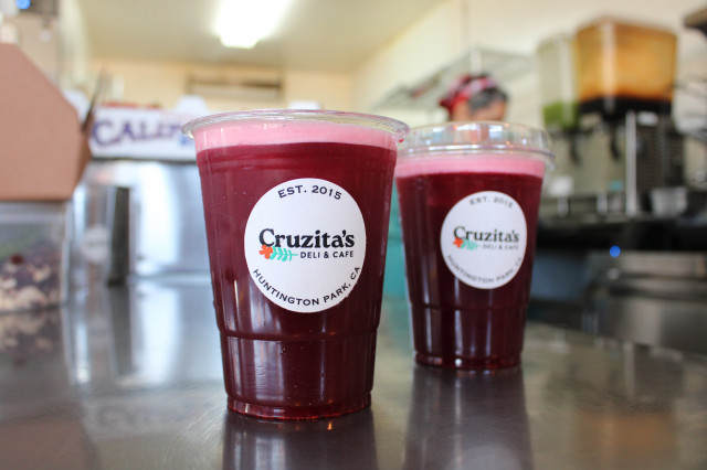 Two clear plastic cups filled with a vibrant, deep red liquid and topped with a light pink foam, featuring the Cruzita's Deli & Cafe logo on a stainless steel counter.