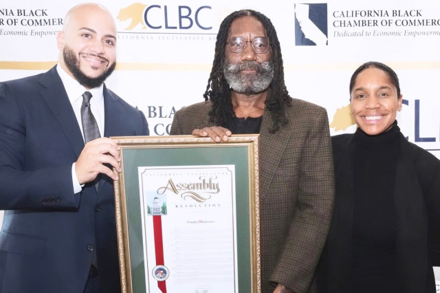 Two men and one women of stand in front of a step and repeat banner. The two men hold between them a certificate of recognition for the restaurant Simply Wholesome