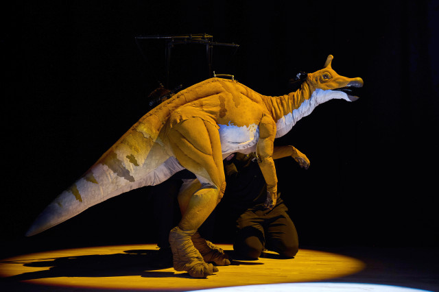 Duck-billed and horned camel and white-colored dinosaur puppet in profile under a yellow spotlight