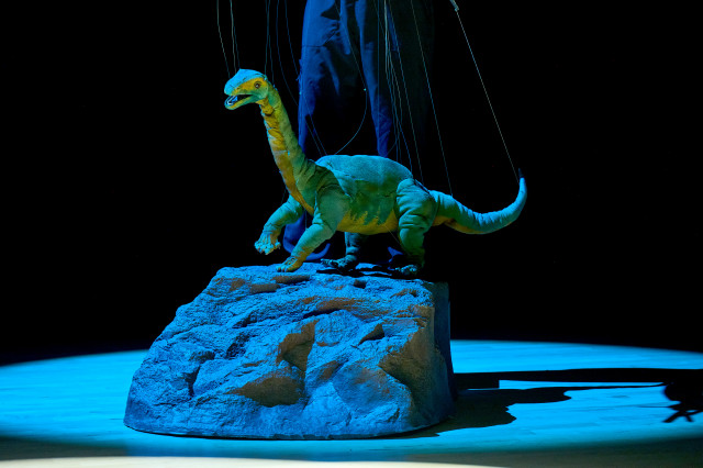 Green long-necked dinosaur puppet, on a faux rock, with clear strings extending upwards and puppeteer's legs behind it