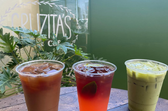 A close up of three cups with colorful aguas frescas, one brownish-red, one red, and one green. On wood table top with green background. 