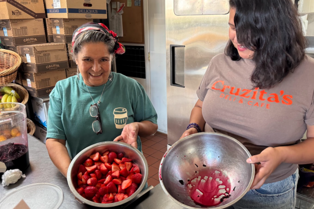 Maricruz, in a teal t-shirt, and Celina, in a brown t-shirt, smiling while holding large stainless steel bowls, one of which is filled with sliced bright red strawberries.