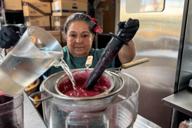 Maricruz wears black gloves and uses a wooden tool to press a thick red fruit pulp through a metal mesh strainer into a large clear container as she pours water over it.