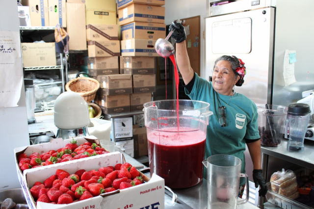Maricruz smiles as she raises a metal ladle high, pouring a stream of bright red liquid into a large transparent container labeled with measurements, with boxes of fresh strawberries in the foreground.
