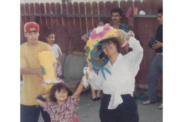 Maricruz and a young Celina hold a broken piñata while Heric, wearing a yellow shirt, stands nearby in an apartment driveway.
