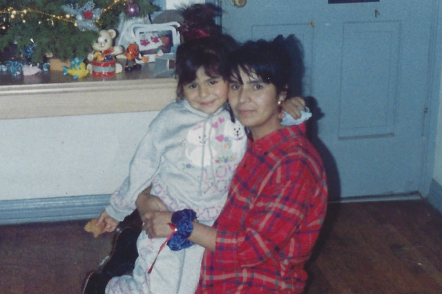A woman in a red plaid shirt holds a young girl in a gray tracksuit in a living room with a decorated Christmas tree.