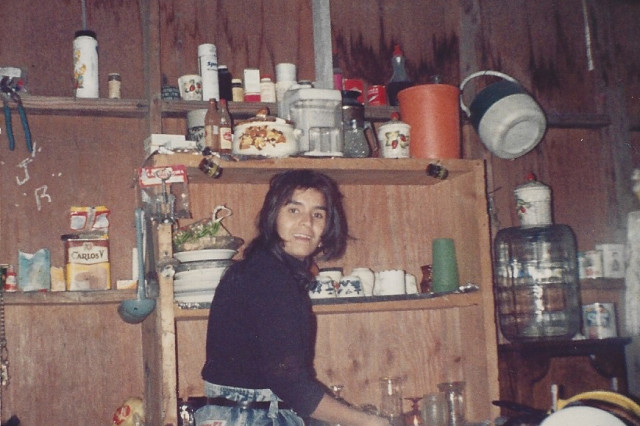 Candid 1990s photo of a young Maricruz smiling in a rustic Tijuana kitchen, standing before wooden shelves packed with pantry items, jars, and cooking utensils.