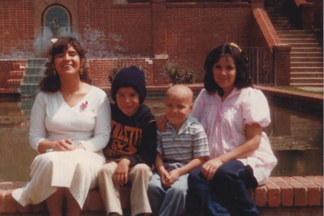 Two women and two young boys, one with a shaved head, sit together on a brick ledge in front of a fountain.