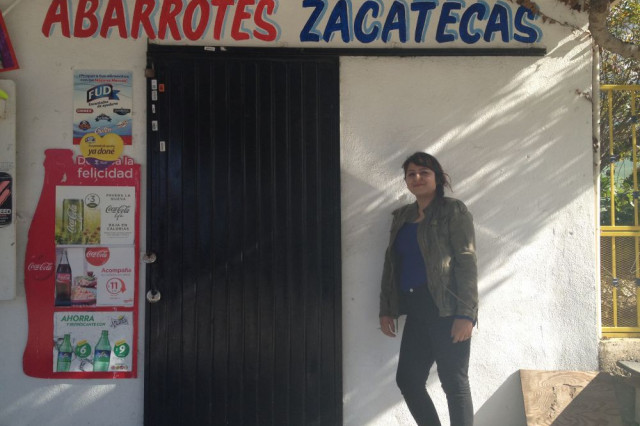 A woman stands outside a small storefront in Tijuana beneath a hand-painted sign that reads “Abarrotes Zacatecas,” with drink advertisements posted beside the door.