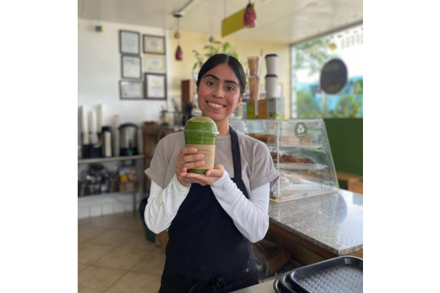 Women in an apron, holding a green smoothy in a plastic cup in a smoothy shop