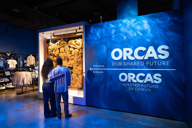 Couple with their arms around each other, facing a mural with Orcas in white capital letters against an ocean background