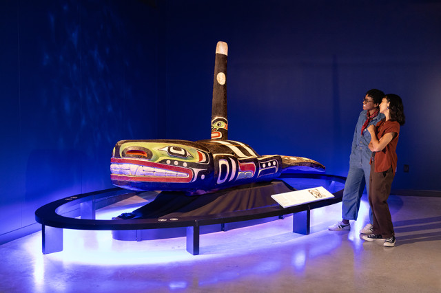Two visitors looking at a wood whale sculpture with symbolic markings