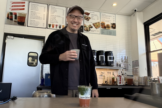 man holding cup and smiling in back ground in foreground Carrot Matcha Latte showing orange, white, and green layers.