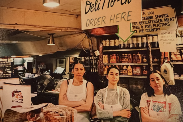 Photograph of three young women standing behind a deli counter with arms crossed over their aprons.