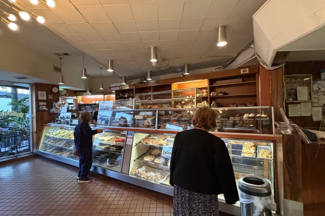 Two older women waiting in line in front of a glass bakery counter with a large array of pastries to choose from.
