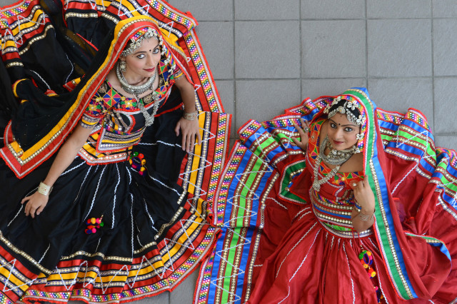 Two women photographed from above, wearing black, red, and multicolored dresses and headdresses, looking up and smiling at the camera