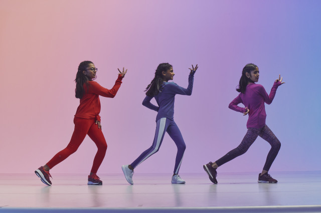Three girls in red, blue, and fuschia in profile with one arm raised and one arm on their waist 