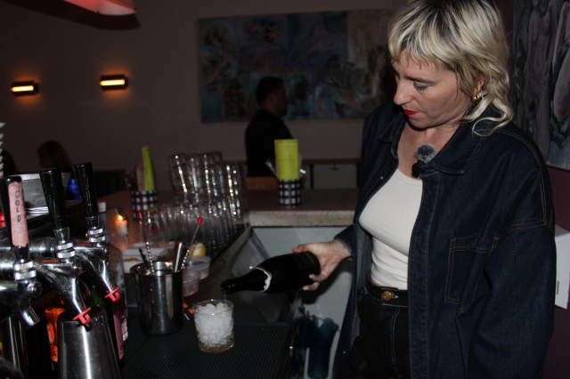 Blond woman pouring a bottle of champagne into an iced hardball glass. 