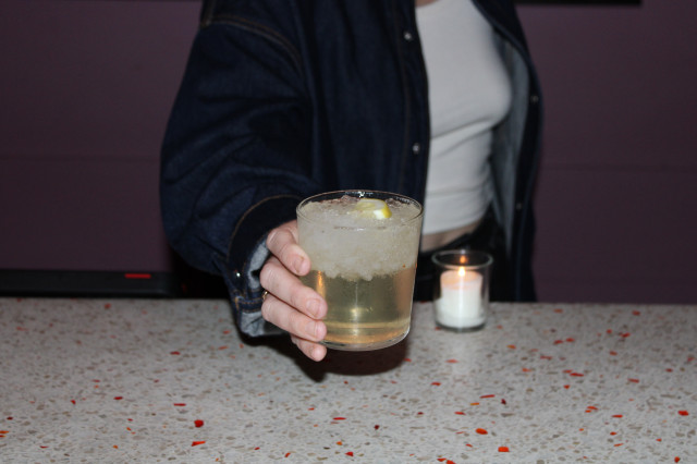 Close-up of woman in white shirt and jean jacket holding an iced cocktail in a highball glass, topped with a slide of lemon. 