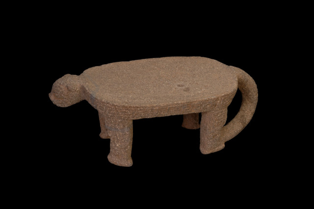 An ancient, textured stone metate (grinding slab) carved in the shape of a four-legged animal, likely a jaguar. It features a flat, oval grinding surface, a small head extending from the left, and a curved tail looping down from the right side, all set against a black background.