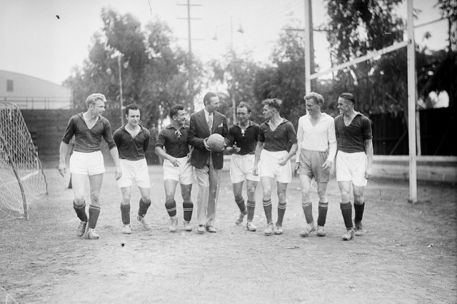 1930s black and white photo of actor Bela Lugosi walking across a soccer field surrounded by seven soccer players in uniform.