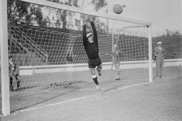 1930s black and white photo of goalie in a tam hat jumping midair to block a ball coming at the net.