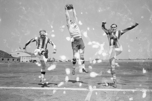 Distressed black and white photo from the 1930s of three soccer players kicking and jumping midair on an air field.