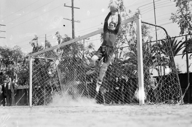 1930s black and white photo of a goalie blocking a ball midair in front of the net.